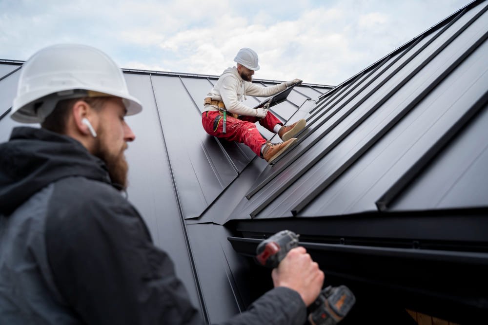 Two construction workers are installing metal roof waterproofing in Singapore. One is seated on the roof, while the other holds a power drill.